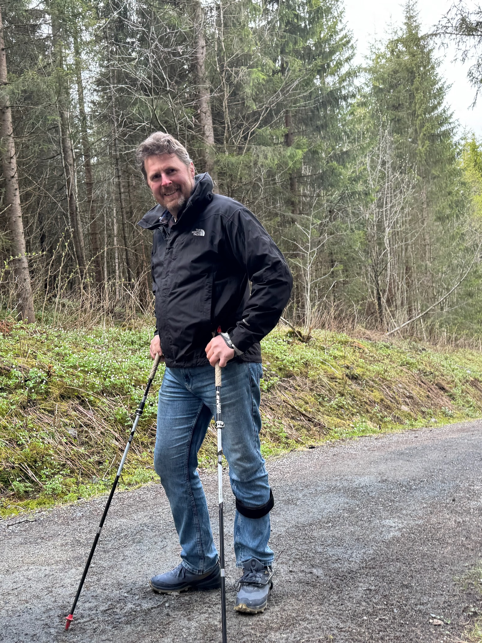 Franz Hölzl lächelnd mit Wanderstöcken auf einem Waldweg zwischen Fichten